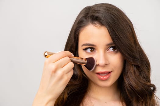 Girl powdering her nose with a brush on the grey background.