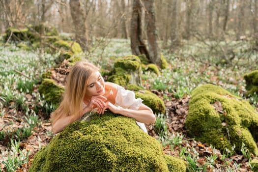 Snowdrops galanthus blond. A girl in a white dress lay down on a stone in the moss in a meadow with snowdrops in a spring forest.