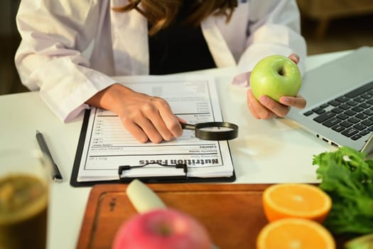 Cropped image of nutritionist holding an apple and prescribing recipe at desk with fresh fruit. Healthcare and diet concept.