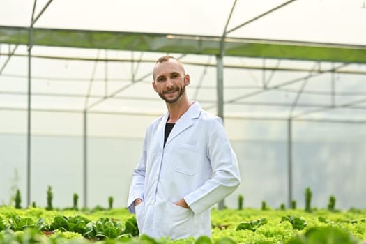 Positive caucasian male plant researcher, geneticist, biologist in white coat standing among vegetable in industrial greenhouse.