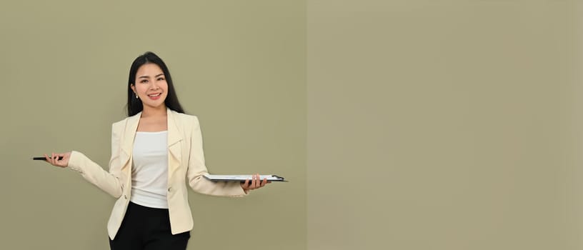 Confident asian businesswoman in stylish suit holding clipboard standing isolated over gray background. Copy space for your text.