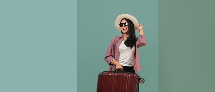 Happy Asian woman wearing summer hat and sunglasses with luggage standing isolated turquoise blue green background.