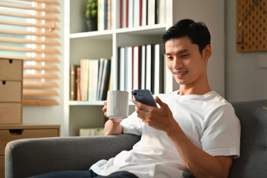 Relaxed asian man drinking coffee and checking social media on his smartphone while sitting on couch at home.