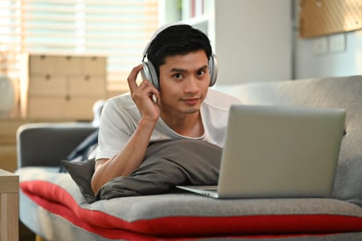 Carefree millennial man wearing headphone and surfing internet or working online on laptop at home.