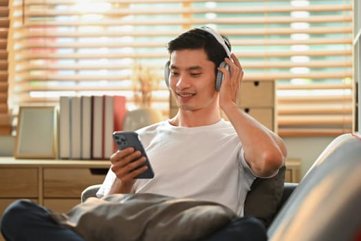 Handsome asian man in casual clothes listening to music in headphone and using smart phone on couch.