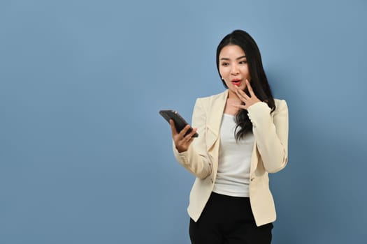 Surprised young Asian woman in stylish suit holding smart phone in hands, standing on blue background.
