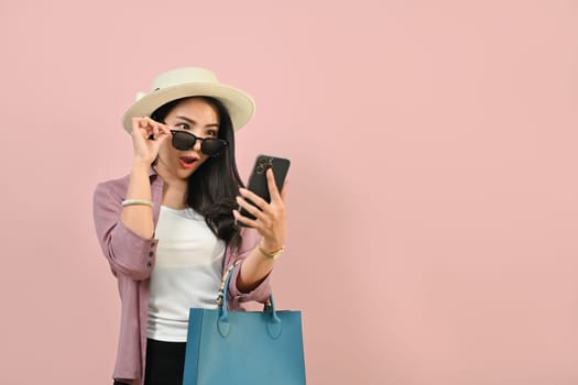 Surprised and amazed woman dressed in summer clothes looking at mobile phone isolated over pink background.