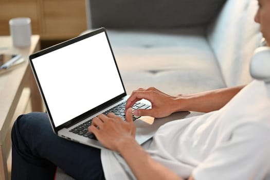 Close up view of casual man browsing internet on laptop, resting on couch in bright living room.