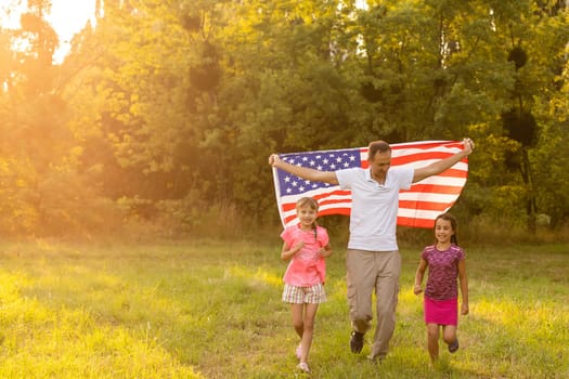 parents and child with American flag are playing with a colorful kite. mother, father and their little daughters celebrate together 4th of July outdoors in foggy day. Independence Day of USA concept