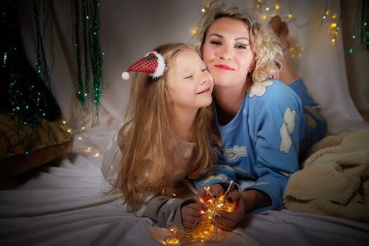 Cute mother and daughter in pajamas lie on soft blanket and having fun in room with Christmas garlands and white background. Tradition of decorating house for holiday. Happy childhood and motherhood