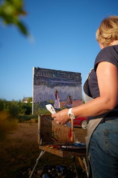 Adult female artist painting picture on the coast of water of a river or lake in nature and trees and greenery in the background