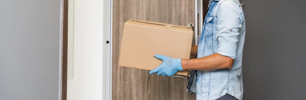 Delivery. Cheerful young courier holding a cardboard box while standing at the entrance of apartment.