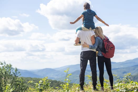 Family standing with arms around on top of mountain, looking at beautiful summer mountain landscape. People enjoying view