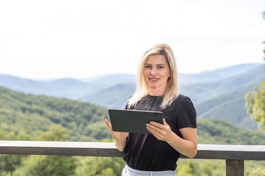 Young woman sits at the terrace with a tablet against beautiful mountain scenery