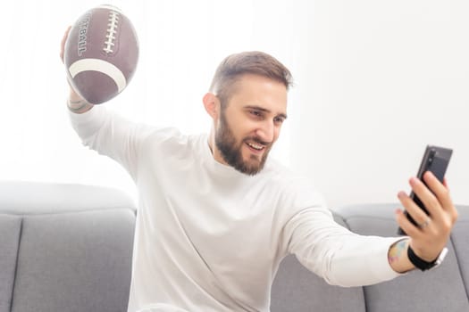 American football fan holding rugby ball and watching match on smartphone.