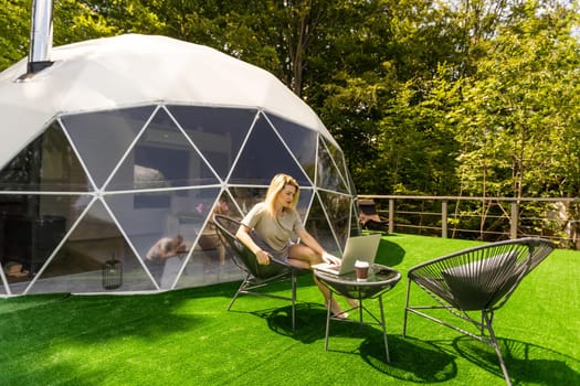 Happy young woman chatting online by using laptop in dome camping. Glamping vacation concept.