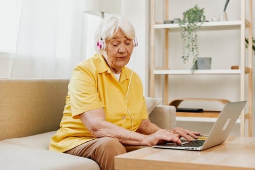 An elderly woman with headphones and a laptop sits on the couch at home and works. High quality photo