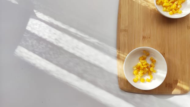 Lot of pieces of canned yellow corn on plate which is on wooden bamboo cutting board on white background. Concept of cooking and delicious healthy food