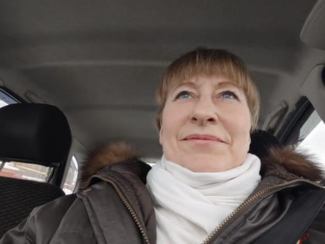 Portrait of adult cute funny blonde woman in warm clothes taking a selfie inside the car and driving it. The concept of pleasant trip in spring, autumn, winter