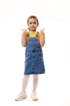 Full-length portrait of Caucasian lovely little girl in casual denim clothes, looking at camera, gesturing with thumbs up, standing over isolated white background. Schoolgirl. Portrait of a smart kid