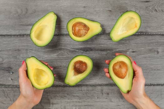 Tabletop view, woman hands holding avocado cut in half, seed visible, more avocados halves on gray wood desk around.