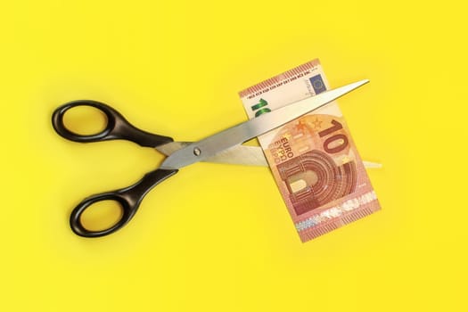 Tabletop view - large scissors cutting ten eur banknote over yellow board. Cut fees concept.