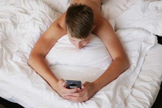 A cute boy lying on a bed with a white bed looks into a smartphone. A boy in shorts lies on a white bed with a phone in his hands, top view.