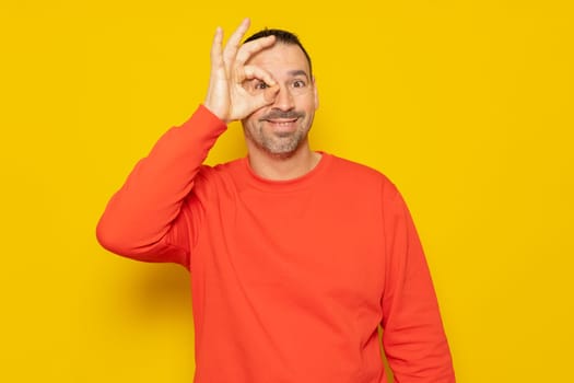 Bearded hispanic man in his 40s wearing a beige turtleneck making an ok gesture looking through fingers, isolated on yellow studio background