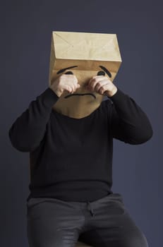 A sad man in a black turtleneck with a bag on his head, with a drawn crying emoticon, cries and wipes away a tear. Emotions and gestures. Vertical frame.