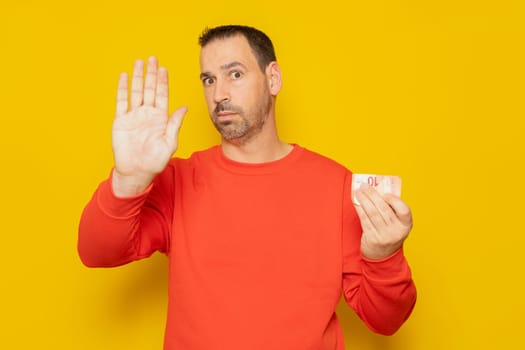 Bearded hispanic man in his 40s wearing a red jumper holding a 10 euro banknote and making the stop sign isolated over yellow background