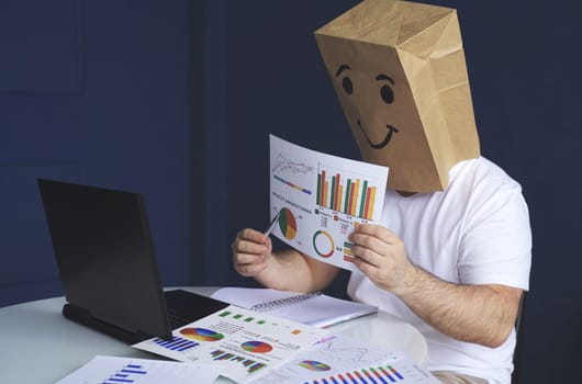 A man is a businessman in a white shirt with a paper bag on his head, with a joyful smiley face drawn, conducts a video conference or training via video link on a laptop. Emotions and gestures.