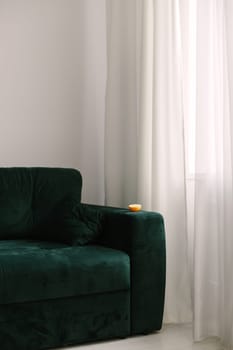 There is an orange on a green sofa by the window. Vertical frame.