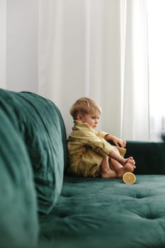 A boy in a yellow bathrobe sits on a green sofa, half an orange lies next to him.