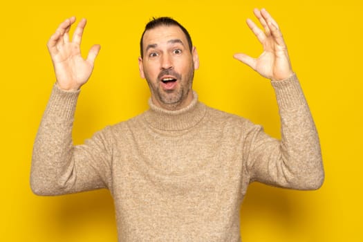 Bearded Hispanic man in his 40s wearing a beige turtleneck raising his arms energetically after receiving a big surprise, isolated on yellow studio background