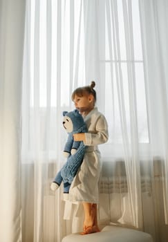 A girl in a dressing gown stands by the window and plays with a knitted toy.