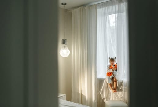 A girl in a bathrobe plays on the windowsill with a knitted lion.