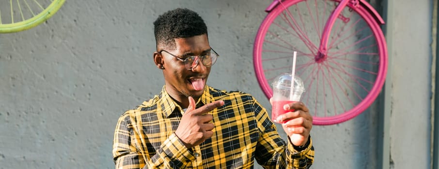 Carefree african american guy in casual wear holding tropical cocktail urban summer background. Cool young man drinking yummy alcohol free beverage on summer vacation