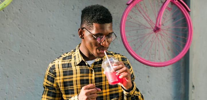Carefree african american guy in casual wear holding tropical cocktail urban summer background. Cool young man drinking yummy alcohol free beverage on summer vacation
