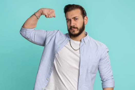 I am strong. Fit sporty lebanese man showing biceps and looking confident, feeling power strength to fight for rights energy to gain success win. Arabian guy isolated on studio blue background indoors