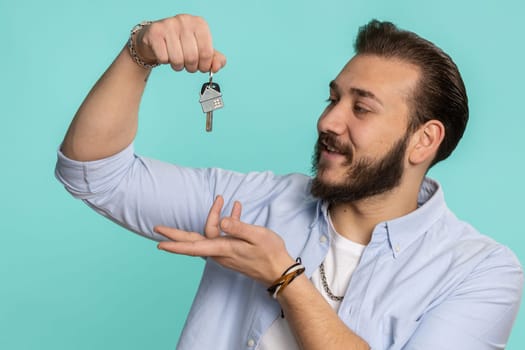 Young lebanese man real estate agent lifting hand showing the keys of new home house apartment, buying or renting property, mortgage loan. Bearded arabian guy isolated on blue studio background