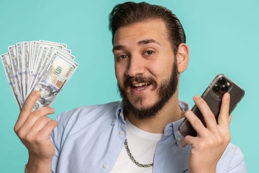 Lebanese man looking smartphone display sincerely rejoicing win, receiving money dollar cash banknotes, success lottery jackpot luck, income salary wage. Arabian guy isolated on studio blue background