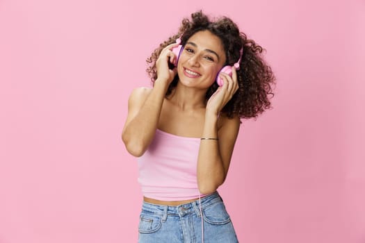 Happy woman wearing headphones with curly hair listening to music and dancing in a pink T-shirt and jeans on a pink background, copy space. High quality photo