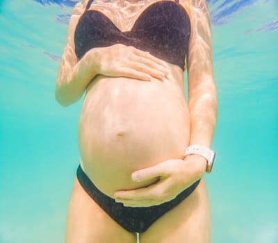 Pregnant woman underwater swimming in tropical sea. Healthy and active pregnancy. Young expecting mom on summer beach vacation before baby birth. Swim holiday and water fun. Travel during pregnancy. Underwater photography.