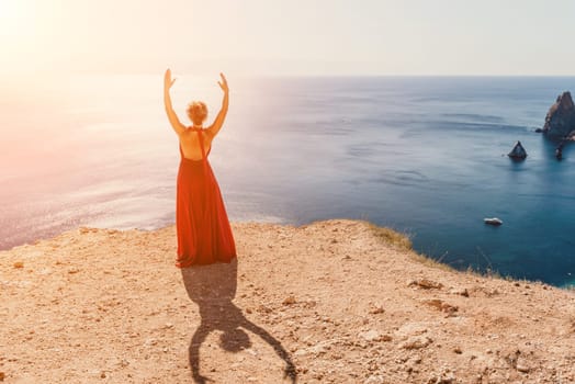 Side view a Young beautiful sensual woman in a red long dress posing on a rock high above the sea during sunrise. Girl on the nature on blue sky background. Fashion photo.