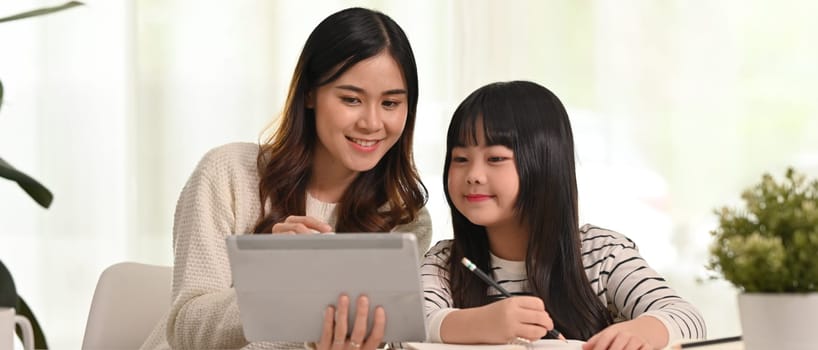 Smiling mother and little daughter sitting at desk in living room studying online and doing assignments together.