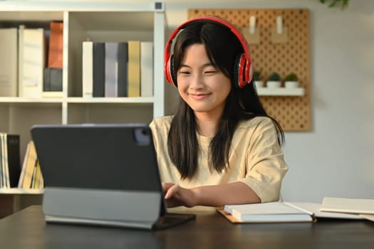 Happy asian girl wearing headphones sitting at table learning online at virtual class on laptop. Distance education concept.
