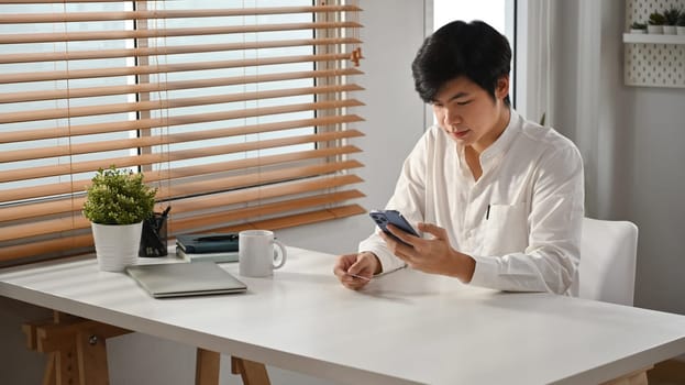 Millennial asian man sitting in modern home office and using smartphone. People, Technology and Communication.