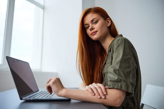 portrait of a cute, smiling red-haired woman sitting at a laptop with her hands folded on the table and looking at the camera. High quality photo