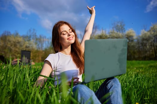 a woman works at a laptop sitting in a field and joyfully raises her hand up. High quality photo