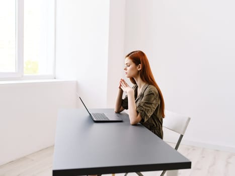 a slender, red-haired woman works at a laptop in a bright office room. High quality photo